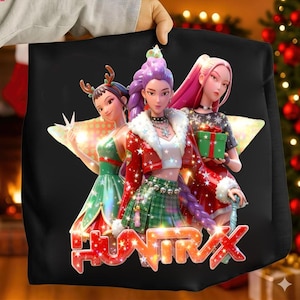 Puede incluir: Sudadera negra con un gráfico de tres mujeres animadas con atuendos festivos. Las mujeres están sobre un fondo en forma de estrella, con la palabra "HUNTRIX" en letras rojas y blancas en la parte inferior. Tema navideño.