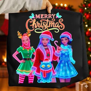 Puede incluir: Sudadera negra con un gráfico de "Merry Christmas". El diseño presenta a tres mujeres de dibujos animados con trajes festivos, incluyendo gorros de Papá Noel y un gato con bufanda. Las mujeres visten trajes verdes, rojos y azules.