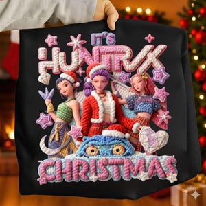 Puede incluir: Sudadera negra con un gráfico colorido que presenta tres figuras con gorros de Papá Noel y el texto "IT'S HUNTRX CHRISTMAS". El diseño incluye estrellas y una criatura azul con gorro de Papá Noel.