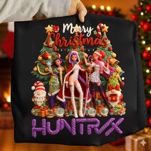 Puede incluir: Sudadera negra con un gráfico navideño. El diseño presenta tres figuras de estilo anime frente a un árbol de Navidad, con las palabras "Merry Christmas" y "HUNTRX" en morado.