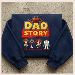 May include: Navy blue sweatshirt featuring the embroidered phrase "DAD STORY" in yellow and red, with a brown cowboy hat above. Below are embroidered cartoon characters and names: Jack, Anna, Sophia, and Lucian.