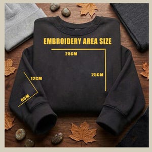 May include: A black sweatshirt with the text "EMBROIDERY AREA SIZE" and dimensions in centimetres. The embroidery area is 25cm x 25cm. The sleeve cuff is 6cm x 12cm. The sweatshirt is displayed on a wooden surface with autumn leaves.