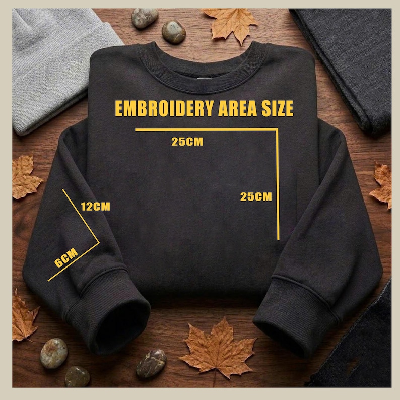 May include: A black sweatshirt with the text "EMBROIDERY AREA SIZE" and dimensions in centimetres. The embroidery area is 25cm x 25cm. The sleeve cuff is 6cm x 12cm. The sweatshirt is displayed on a wooden surface with autumn leaves.