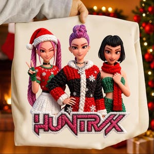 Può includere: Una borsa tote color crema con una grafica di tre figure femminili stilizzate in abiti invernali. Le figure indossano maglioni, cappelli e sciarpe rossi e verdi. La parola "HUNTRAX" è visualizzata in un carattere stilizzato nella parte inferiore della borsa.