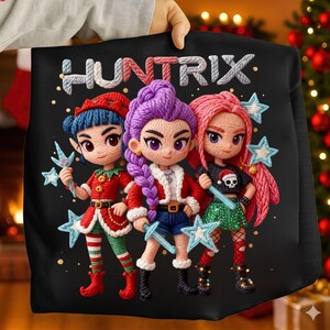 Puede incluir: Sudadera negra con la palabra "HUNTRIX" sobre tres personajes de dibujos animados. Los personajes tienen pelo azul, morado y rosa, y visten trajes festivos con varitas en forma de estrella. El fondo es negro con pequeños puntos dorados.