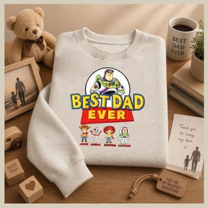 May include: A light gray sweatshirt with the text "BEST DAD EVER" in yellow and red, featuring a Buzz Lightyear graphic and cartoon characters. Below the text are the names "JACK ANNA SOPHIA LUCIAN".