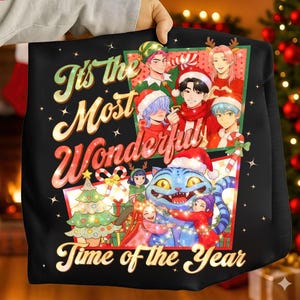 Puede incluir: Sudadera negra con un gráfico colorido que presenta personajes de estilo anime, un gato azul con un gorro de Papá Noel y el texto "It's the Most Wonderful Time of the Year". El diseño incluye elementos navideños.