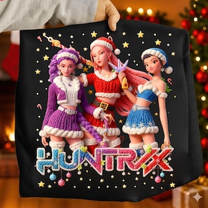 May include: Black sweatshirt featuring three stylized figures in Christmas outfits, surrounded by gold stars. The figures are in purple, red, and blue outfits, with the word "HUNTRAX" in colorful letters at the bottom. The sweatshirt is held by a person.