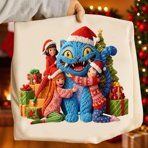 Puede incluir: Un cojín navideño con un gato azul con gorro de Papá Noel, rodeado de tres niños con jerséis y gorros festivos. El cojín también incluye regalos envueltos y un árbol de Navidad, todo sobre un fondo crema.