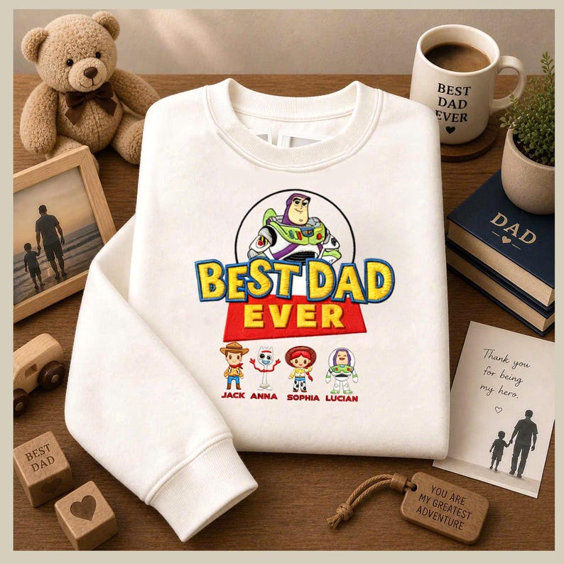 May include: White sweatshirt with the text "BEST DAD EVER" and Toy Story characters. The sweatshirt has a crew neck and long sleeves. A mug with "BEST DAD EVER" is also visible, along with a teddy bear and a wooden block.