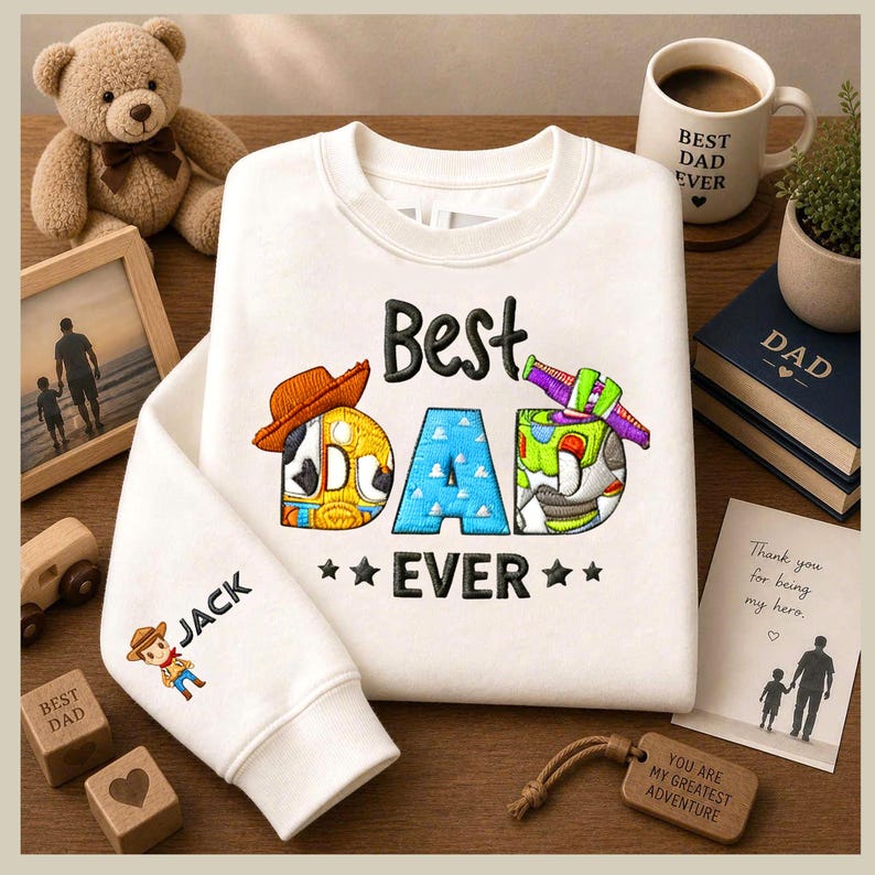May include: Cream-coloured sweatshirt with the embroidered text "Best Dad Ever". The sleeve has the name "Jack" and a cartoon cowboy. A mug with "Best Dad Ever" is also visible, along with a wooden tag.