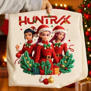Puede incluir: Un cojín crema con un diseño festivo. La palabra "HUNTRX" está en rojo. La imagen muestra a tres mujeres con trajes navideños rojos, cada una con un gorro de Papá Noel. El diseño incluye regalos y adornos navideños.