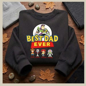 May include: Black sweatshirt with the text "BEST DAD EVER" and a graphic of Buzz Lightyear. Below the text are cartoon characters and names. The sweatshirt is surrounded by fall leaves and small stones.