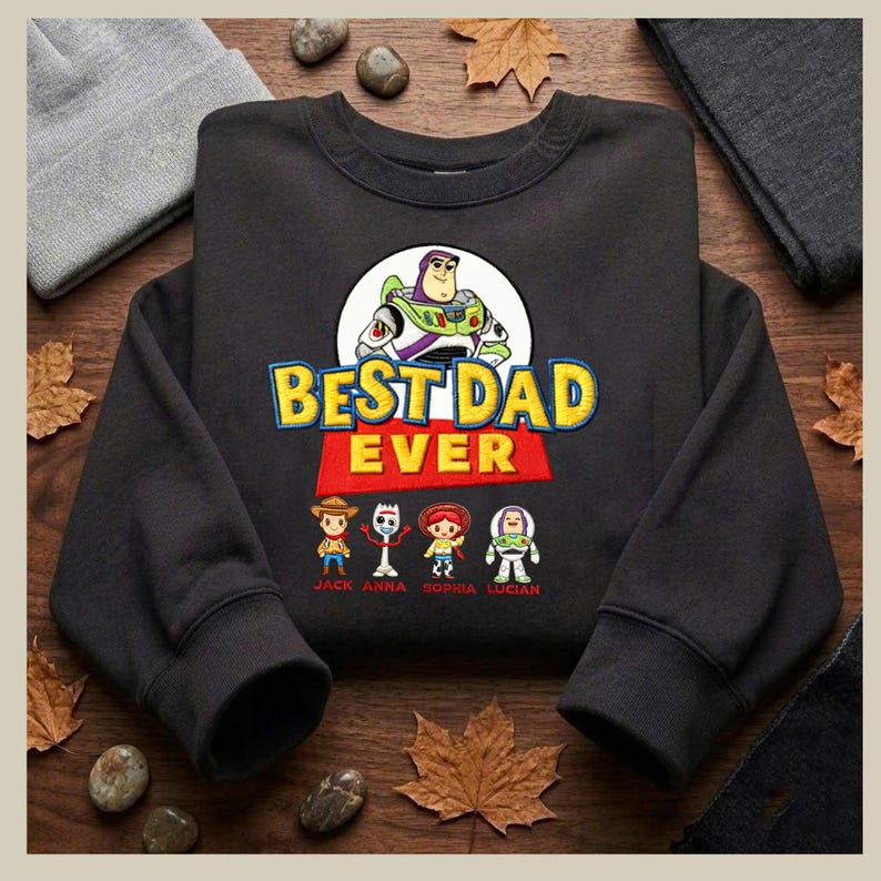 May include: Black sweatshirt with the text "BEST DAD EVER" and a graphic of Buzz Lightyear. Below the text are cartoon characters and names. The sweatshirt is surrounded by fall leaves and small stones.