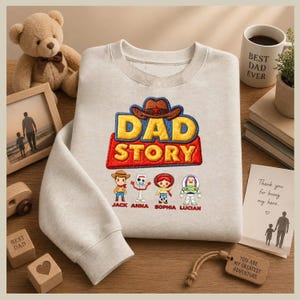 May include: A light grey sweatshirt with the embroidered text "DAD STORY" in yellow and red, featuring cartoon characters. Below the text are the names "JACK ANNA SOPHIA LUCIAN". A teddy bear, framed photo, and wooden blocks are also visible.