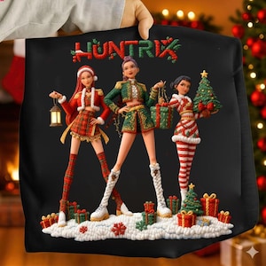Puede incluir: Sudadera negra con la palabra "HUNTRIX" bordada en rojo y verde. El diseño presenta tres figuras estilizadas con atuendos festivos, sosteniendo regalos y un árbol de Navidad. Las figuras están sobre una base nevada con cajas de regalo.