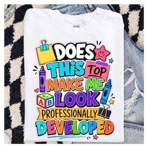 May include: A white t-shirt featuring a colorful graphic with school-themed illustrations and the text: DOES THIS TOP MAKE ME LOOK PROFESSIONALLY DEVELOPED. The design includes a yellow sticky note, a pink note, crayons, a block, and binder clips.