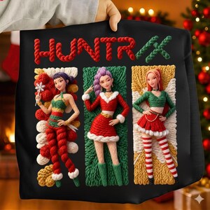 May include: Black sweatshirt with the word "HUNTRX" in red and green rope-like letters. The sweatshirt features three stylized female figures in Christmas-themed outfits, each in a different color scheme.