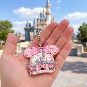 May include: Pink Disney Girl keychain featuring a castle design with a bow and heart accents. The keychain has a silver chain and a pink and white color scheme. The background shows a castle and blue sky.