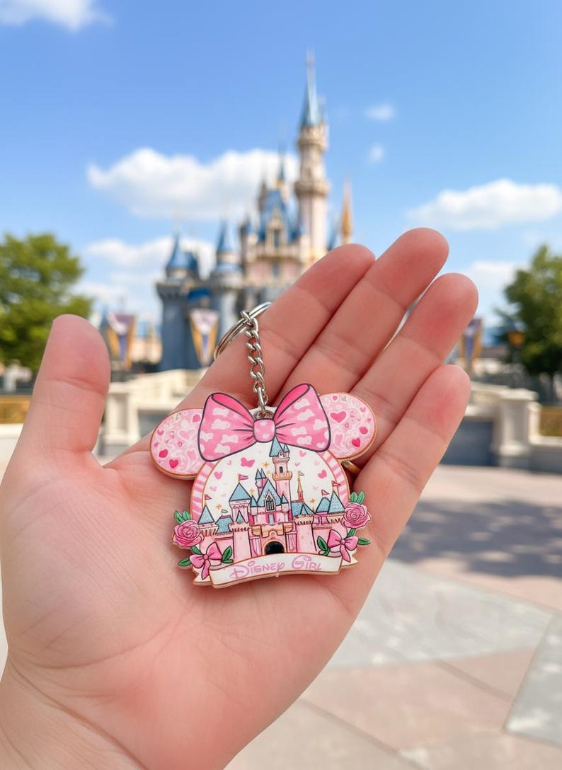 May include: Pink Disney Girl keychain featuring a castle design with a bow and heart accents. The keychain has a silver chain and a pink and white color scheme. The background shows a castle and blue sky.