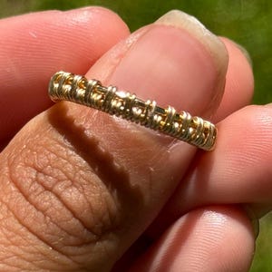 May include: A gold-toned ring with a textured design. The ring appears to be made of intertwined metal, creating a woven or coiled pattern. The ring is held between fingers against a blurred green background.