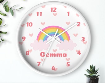 May include: White round clock with pink hearts, a rainbow, and clouds. The clock face has black numbers and hands. The name "Gemma" is written in pink below the rainbow.