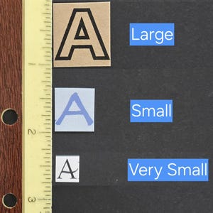 May include: Three square paper cutouts featuring the letter A in different sizes and styles, labeled Large, Small, and Very Small. A yellow ruler with inch markings is positioned vertically on the left, showing the largest A is approximately 1 inch tall.