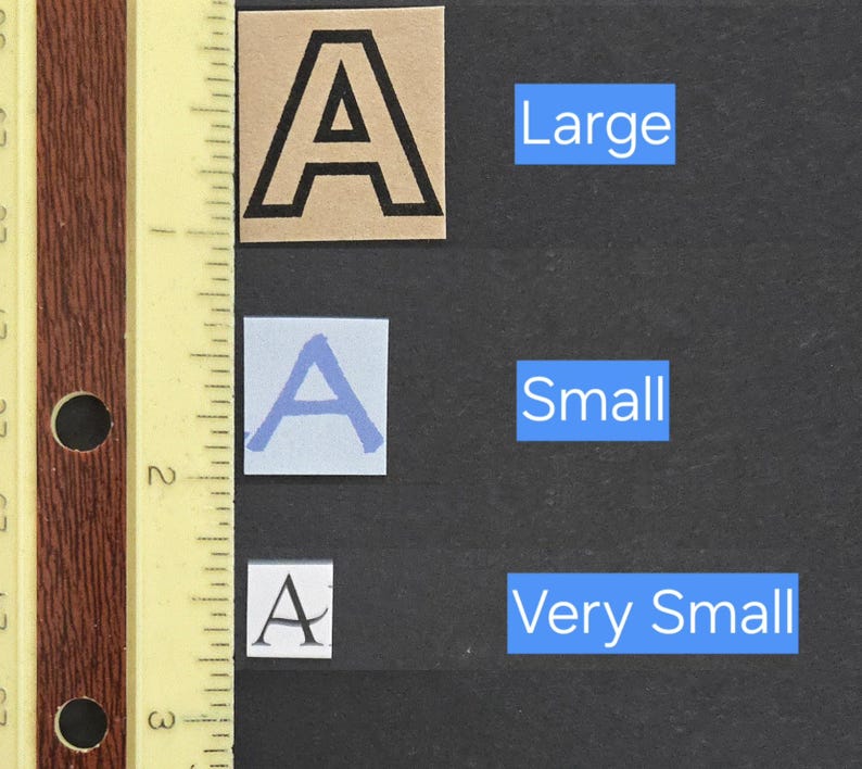 May include: Three square paper cutouts featuring the letter A in different sizes and styles, labeled Large, Small, and Very Small. A yellow ruler with inch markings is positioned vertically on the left, showing the largest A is approximately 1 inch tall.