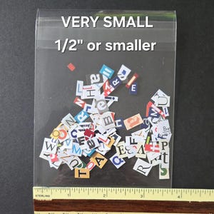 May include: A clear plastic bag containing a collection of small, multicolored paper cutouts of individual letters and characters. The text on the bag reads VERY SMALL 1/2" or smaller. A yellow ruler is positioned at the bottom for scale.