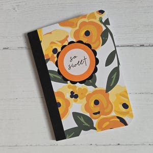 May include: A floral notebook with a black spine. The cover features a pattern of yellow and orange flowers with green leaves on a white background. A circular orange sticker with the words "so sweet" is on the cover.