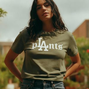 Puede incluir: Camiseta verde oliva con la palabra "pLAnts" en blanco, con una fuente estilizada. La modelo lleva vaqueros azules. El fondo incluye plantas en macetas y un edificio.
