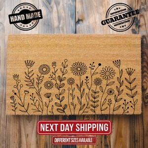 May include: A rectangular doormat made of natural fibers, featuring a black floral design along the bottom edge. The mat has the words "HAND MADE" and "SATISFACTION GUARANTEE" stamped on it. The text "NEXT DAY SHIPPING" and "DIFFERENT SIZES AVAILABLE" are also present.