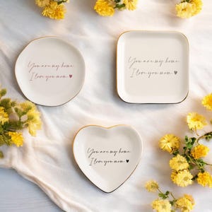 May include: Three white ceramic trinket dishes with gold trim. Each dish has a different shape: round, square, and heart. The dishes have the text "You are my home. I love you mom." printed on them.