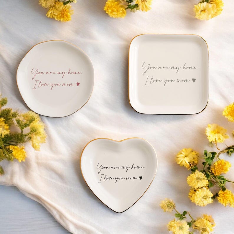 May include: Three white ceramic trinket dishes with gold trim. Each dish has a different shape: round, square, and heart. The dishes have the text "You are my home. I love you mom." printed on them.