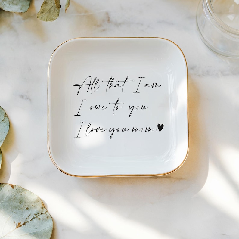 May include: White ceramic trinket dish with a gold rim. The dish has the text "All that I am I owe to you I love you mom." written in black cursive with a small black heart.