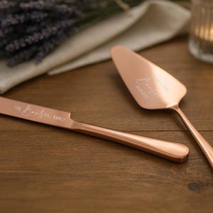 May include: Rose gold cake knife and server set with engraved names and date. The knife reads "The Booth Family". The server reads "Jennifer 06.05.2023". The set is on a wooden surface with candles and lavender.