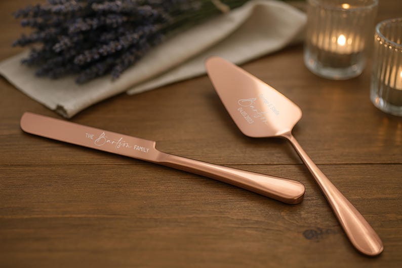 May include: Rose gold cake knife and server set with engraved names and date. The knife reads "The Booth Family". The server reads "Jennifer 06.05.2023". The set is on a wooden surface with candles and lavender.