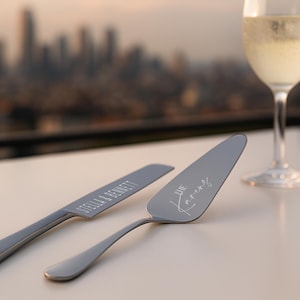 May include: A silver cake knife and server set with engraved names and "The Karens" in script. Two champagne flutes with bubbly liquid are in the background, with a city skyline out of focus.