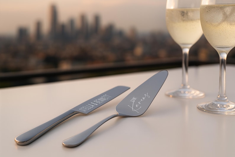 May include: A silver cake knife and server set with engraved names and "The Karens" in script. Two champagne flutes with bubbly liquid are in the background, with a city skyline out of focus.