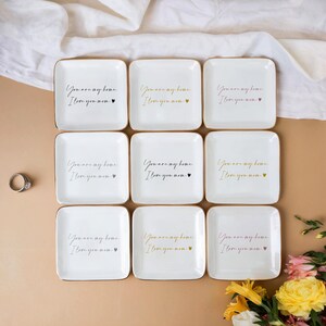 May include: Nine white ceramic trinket dishes with gold trim and the text "You are my home, I love you now" in black script. Each dish has a small gold heart.