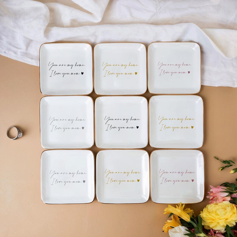 May include: Nine white ceramic trinket dishes with gold trim and the text "You are my home, I love you now" in black script. Each dish has a small gold heart.