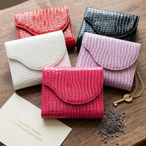 May include: A collection of five small wallets in red, black, white, lavender, and hot pink, each featuring a glossy crocodile-embossed texture and a curved flap closure. A gold-toned key on a chain and a card reading Handcrafted - Premium Croc-Embossed Vegan Leather are included.