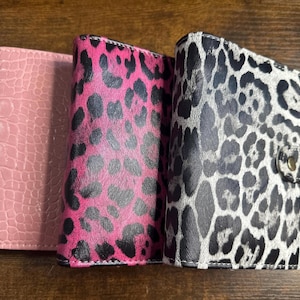 May include: Three stylish wallets in a row. The first is a light pink with a crocodile skin texture. The second is hot pink with black leopard spots. The third is black and white leopard print with a snap closure.