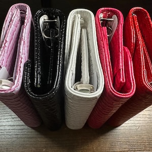 Può includere: Una collezione di cinque raccoglitori ad anelli in vari colori, tra cui rosa, nero, bianco e rosso. I raccoglitori hanno un motivo strutturato, simile alla pelle di serpente, e meccanismi ad anelli in metallo argentato. I raccoglitori sono disposti uno accanto all'altro.