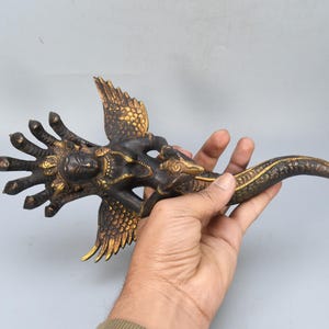 May include: A bronze-colored sculpture of a mythical creature with multiple arms, wings, and a serpent-like tail. The sculpture has gold accents and is held in a person's hand.