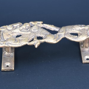 Peut inclure: Une poignée de porte en laiton présentant un motif de dragon sculpté et détaillé. La poignée possède un corps allongé et serpentin avec des écailles texturées et deux plaques de fixation rectangulaires à chaque extrémité, percées d'un trou.