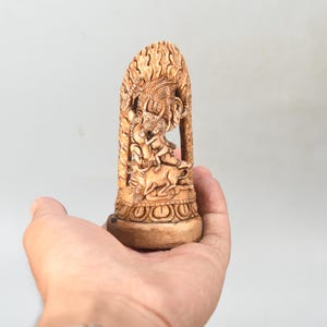 May include: A carved wooden sculpture of a deity riding an animal. The sculpture is light brown with intricate details, held in a hand against a neutral background. The sculpture has a detailed base.