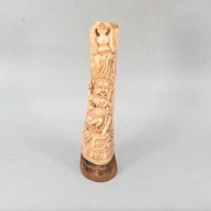 May include: A carved, off-white sculpture of a deity, possibly religious, with intricate details. The sculpture is mounted on a dark brown wooden base. The carvings depict figures and patterns, suggesting a cultural or historical significance. The background is a plain, light gray.