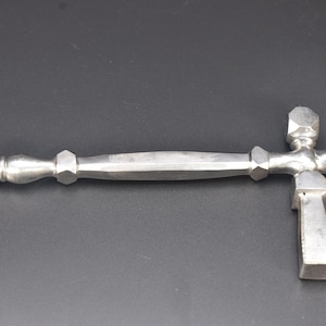 May include: A silver-toned metal tool featuring a long, slender handle with faceted, hexagonal decorative accents and a pointed tip. The opposite end includes a cross-shaped head with a flat, rectangular base extending downward from the main shaft.