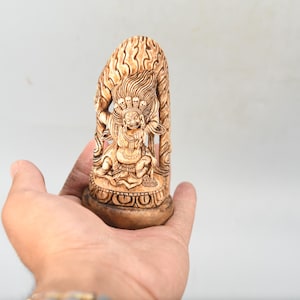 May include: A detailed, carved wooden sculpture of a seated deity with multiple heads and arms. The sculpture is light brown and rests on a round base. The figure is held in a hand against a white background.